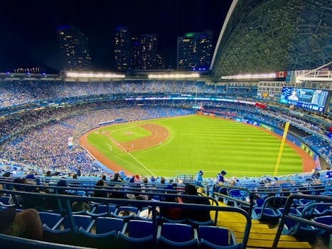 Aerial view of a night baseball game in a packed stadium with city skyline.